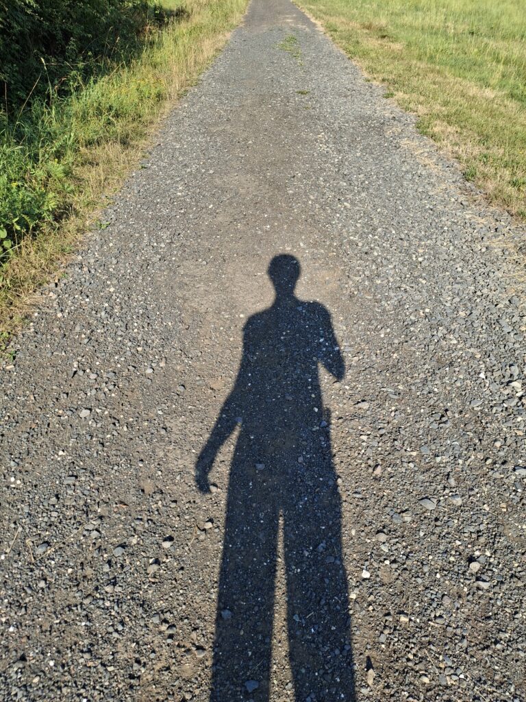 Langer Schatten einer Person auf einem sonnigen Feldweg zwischen Wiesen und Bäumen.
