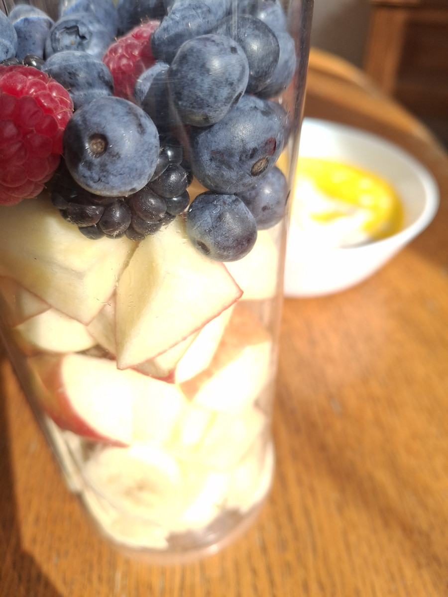 Frische Zutaten für einen Frühstücks-Smoothie: Apfel, Banane, Heidelbeeren und Himbeeren im Mixerbecher im warmen Morgenlicht.