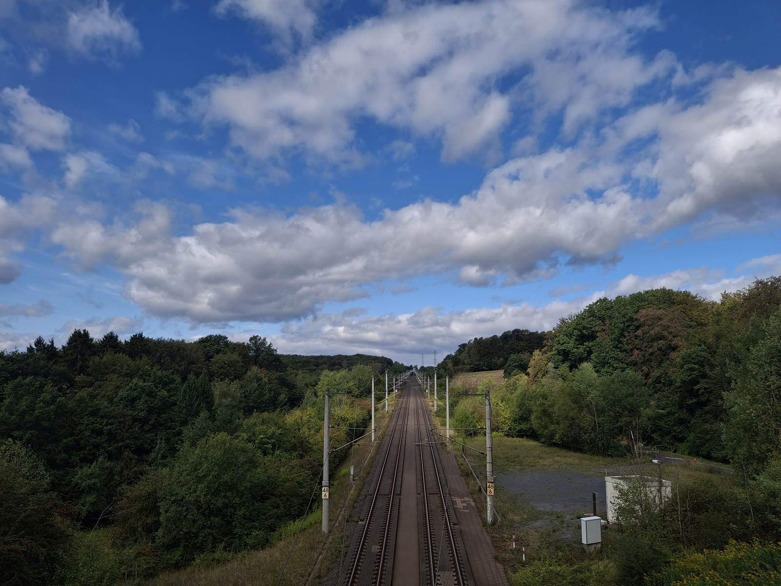 Bahnstrecke, die zwischen grünen Bäumen unter einem weiten Wolkenhimmel in die Ferne führt