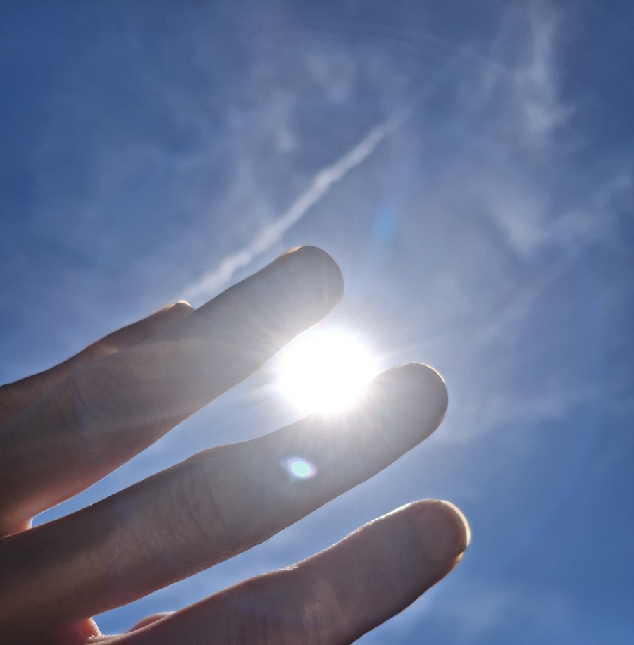 Eine Hand wird vor einen strahlend blauen Himmel gehalten, während die Sonne zwischen zwei Fingern hindurchscheint.