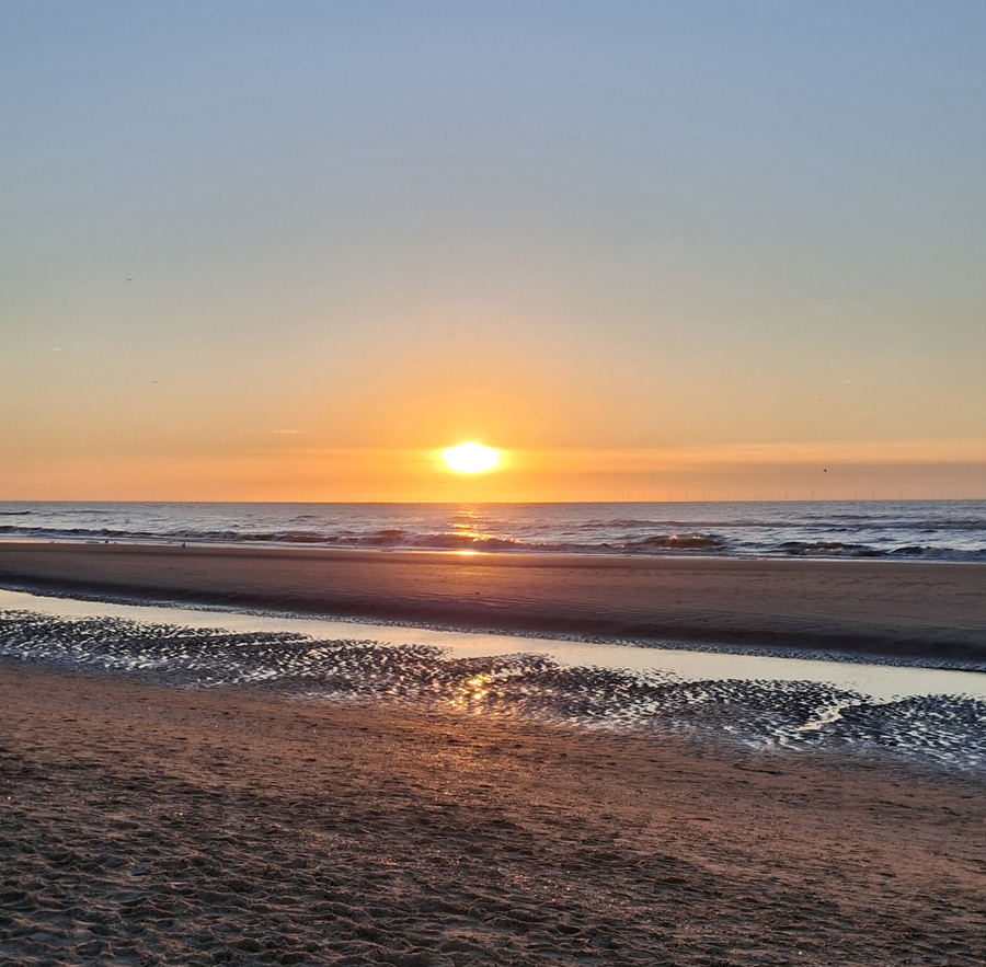 Sonnenuntergang über dem Meer mit sanften Wellen und ruhigem Sandstrand in warmem Abendlicht.