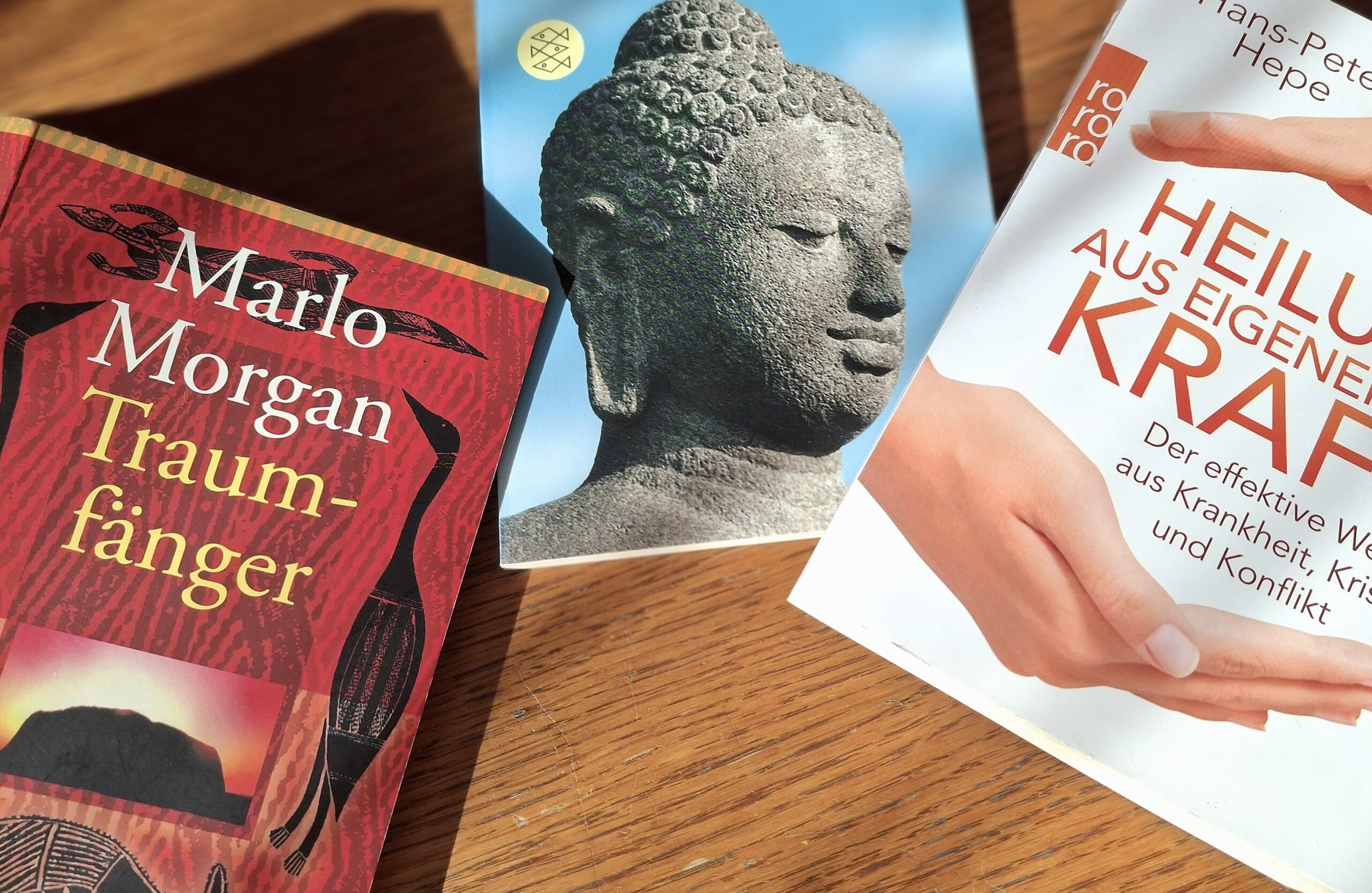 Drei Bücher auf einem Holztisch, darunter „Grundgedanken des Buddhismus“ und „Heilung aus eigener Kraft“